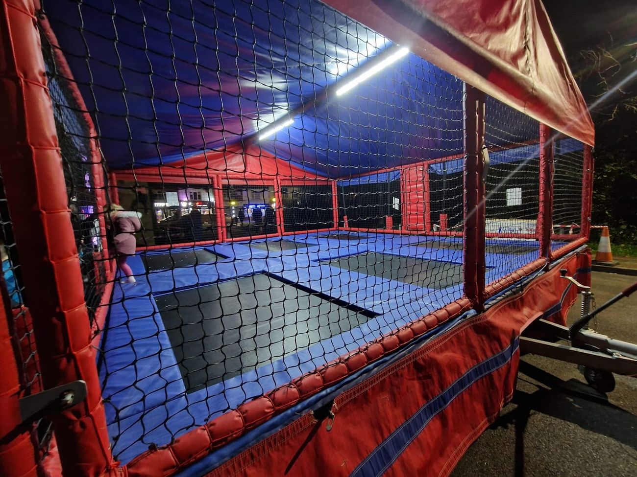 The trampoline set lit from underneath on a town street at night, with the tow hitch in the foreground and lit shopfronts behind