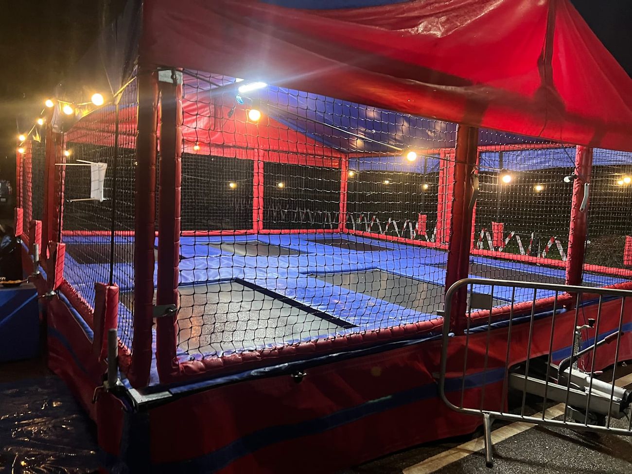 String lights running along the trampoline set's netting and canopy against a dark sky, with crowd barriers in front