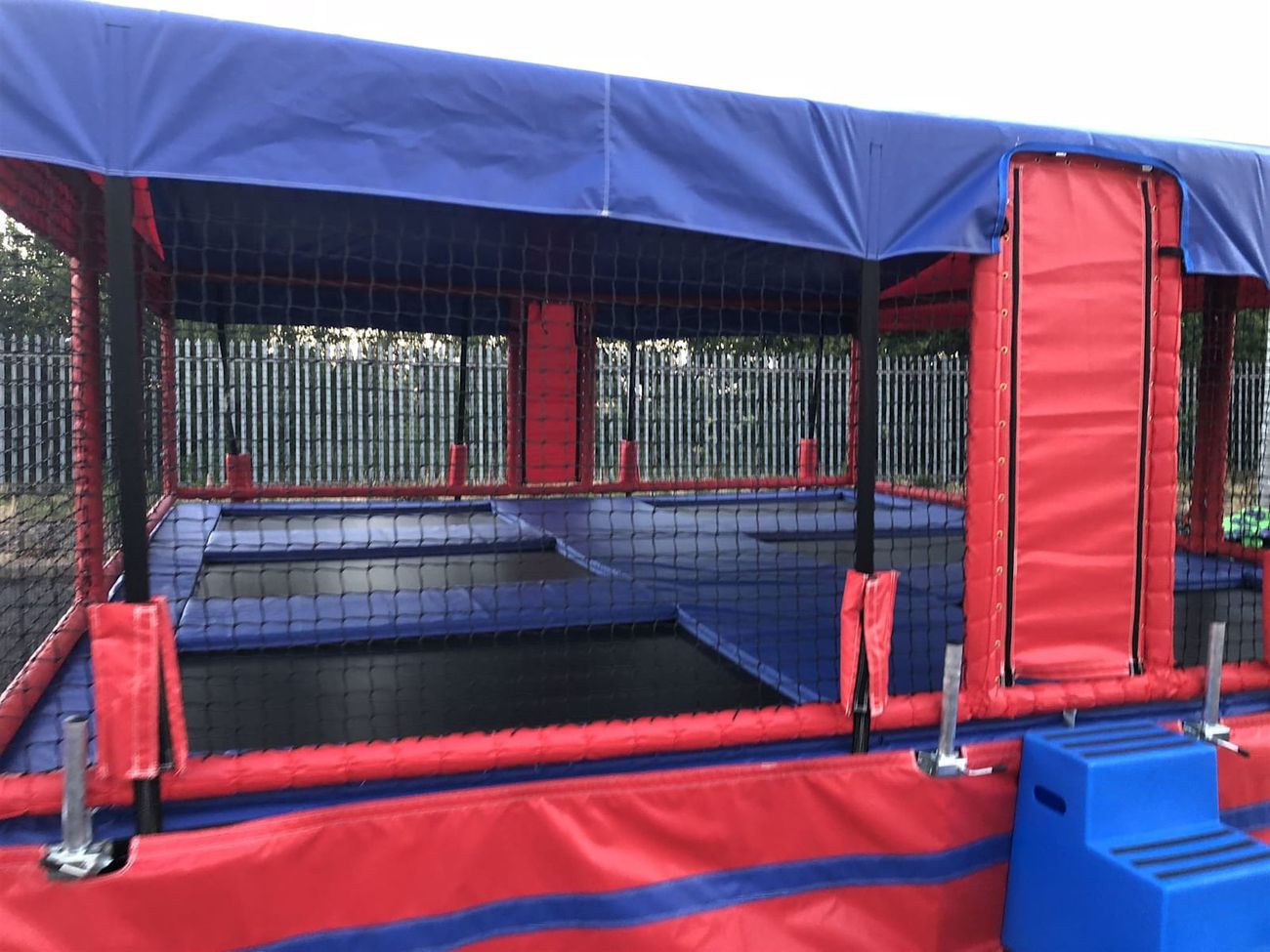 The trampoline beds set into their padded blue frame behind the black safety netting, with the red padded door flap