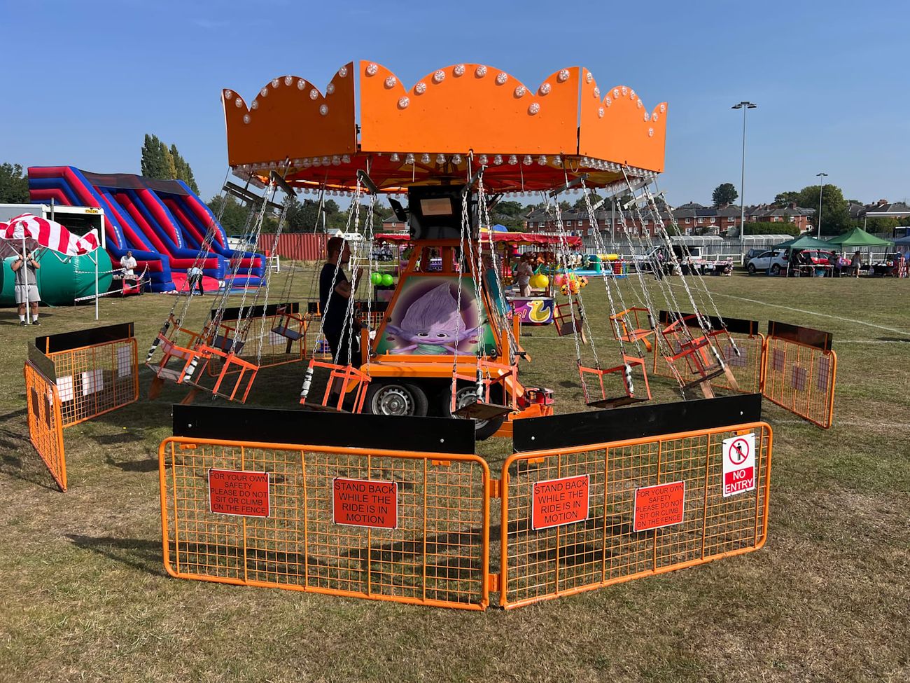 Juvenile Chair-O-Planes on a grass event field, with its orange scalloped canopy, hanging swing seats and character-decorated centre on a trailer base
