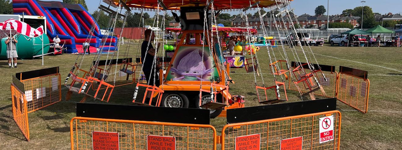 Juvenile Chair-O-Planes on a grass event field, with its orange scalloped canopy, hanging swing seats and character-decorated centre on a trailer base