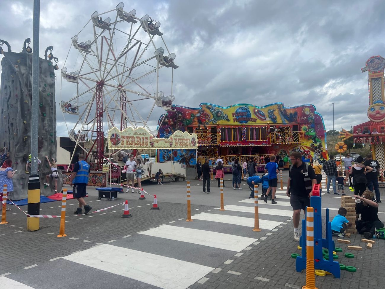 Ferris wheel operating within a larger public funfair beside a climbing wall and colourful funhouse