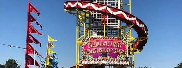 Square-tower helter skelter on a grass field, with the staircase visible inside the mesh tower, a red and white spiral slide and a gold Helter Skelter sign on a pink frontboard