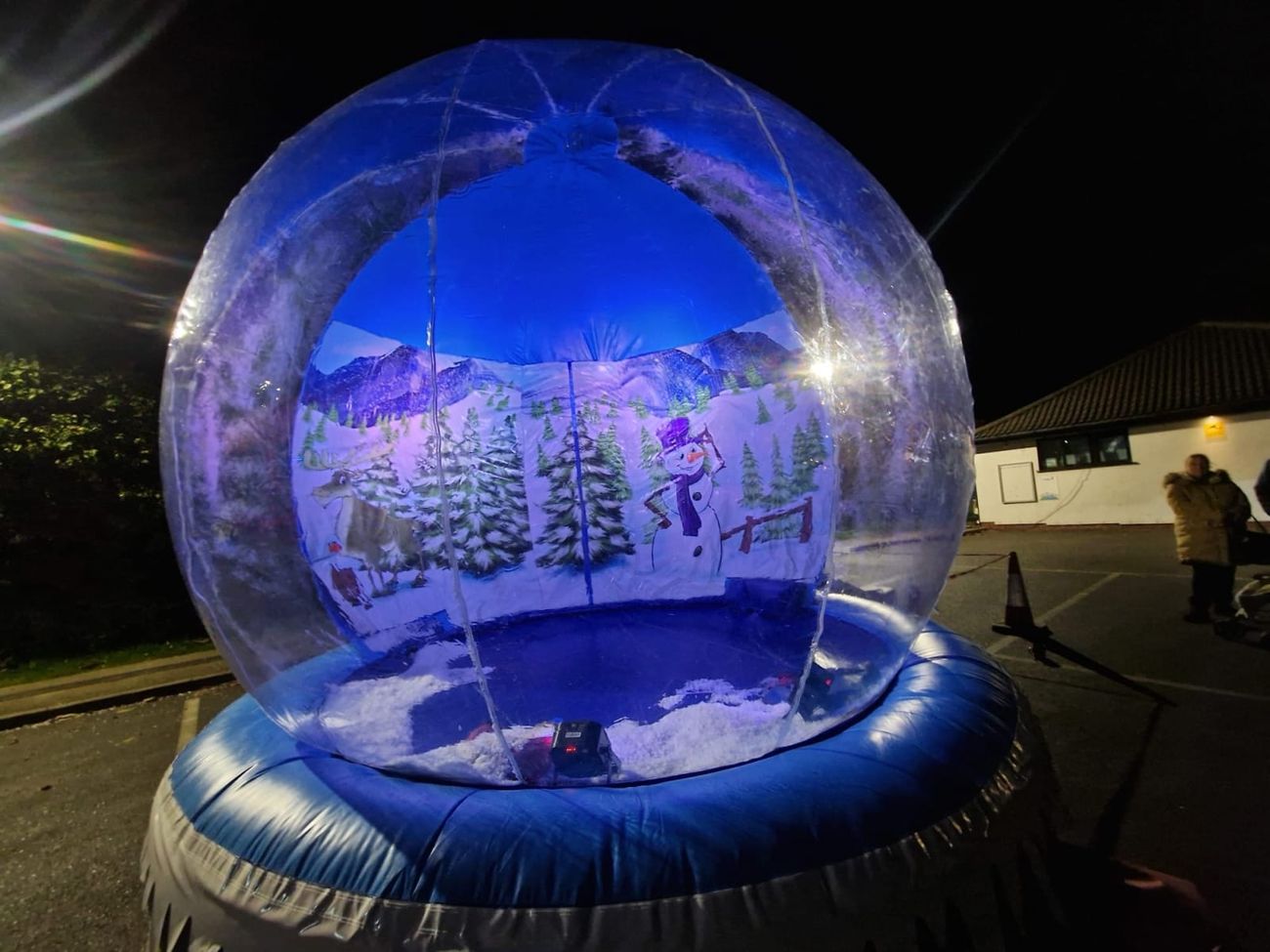 The giant snow globe lit with blue interior lighting at night, the painted winter backdrop visible through the clear dome