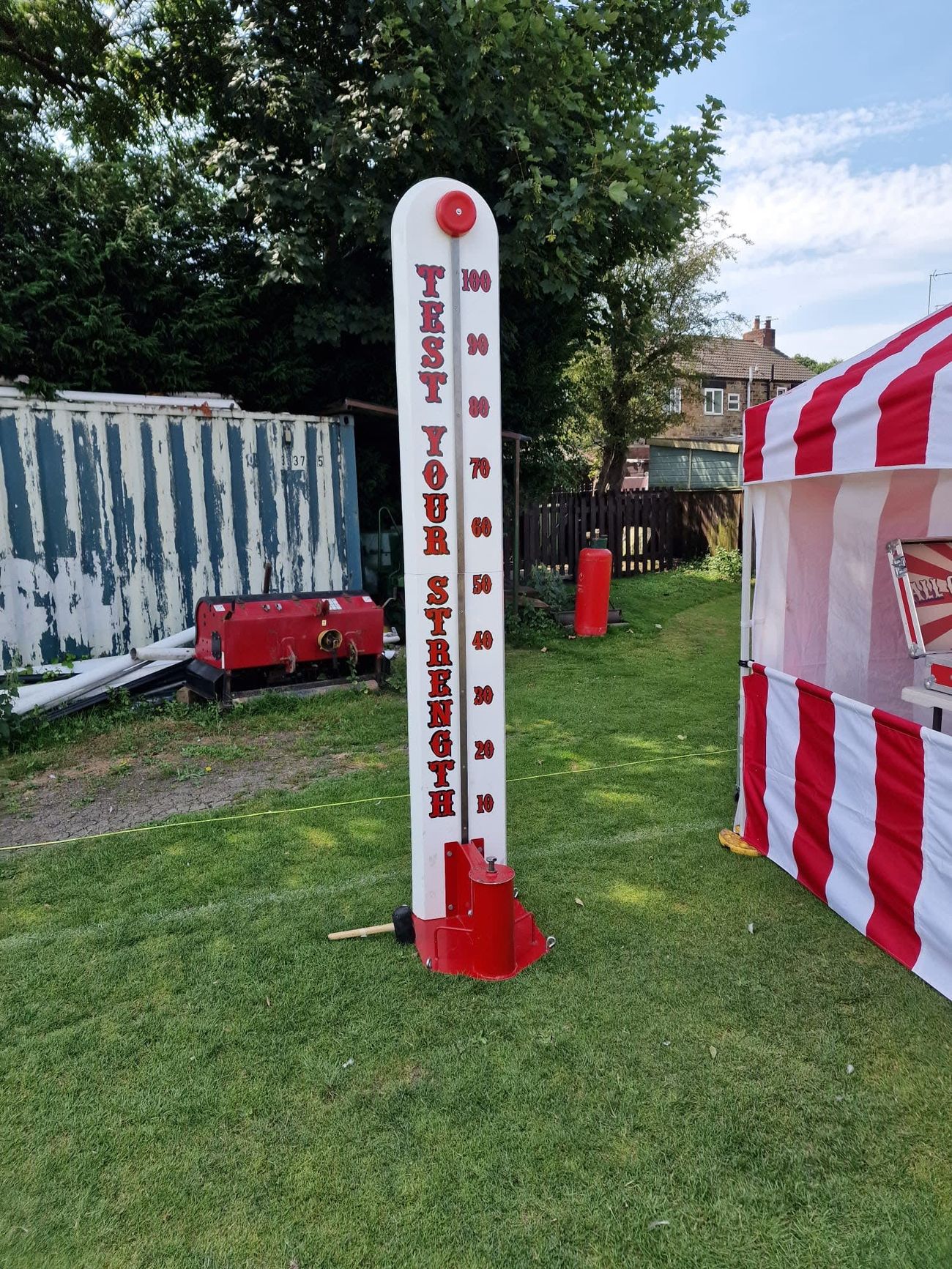 High Striker test-your-strength tower on grass, with its 10-100 scale, mallet on the ground and red bell at the top