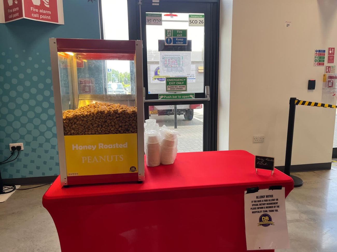Glazed heated nut cabinet on a red-covered table, with its Honey Roasted Peanuts panel, paper cups and chalkboard sign