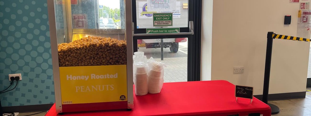 Glazed heated nut cabinet on a red-covered table, with its Honey Roasted Peanuts panel, paper cups stacked beside it and a chalkboard sign
