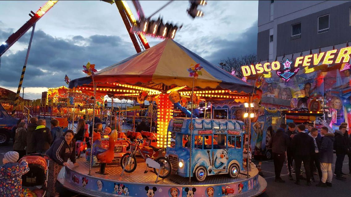 The juvenile toy set roundabout at dusk, with character-painted vehicles on the platform under a red, gold and blue octagonal canopy edged with bulbs