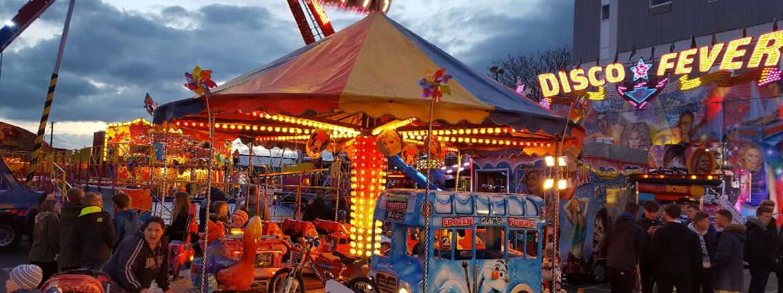 The juvenile toy set roundabout at dusk, with character-painted vehicles on the platform under a red, gold and blue octagonal canopy edged with bulbs