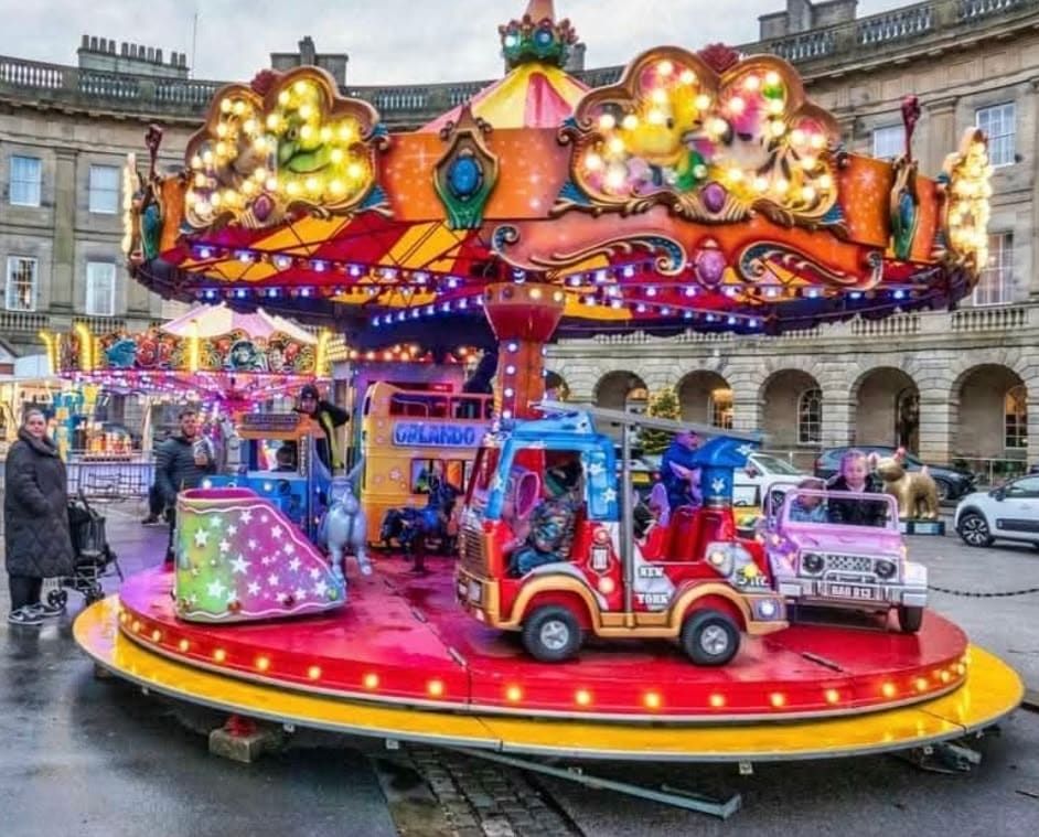 The large juvenile toy set roundabout on a wet paved town square, with the yellow Orlando school bus, red fire engine and pink jeep on the bulb-ringed deck under the orange glitter canopy