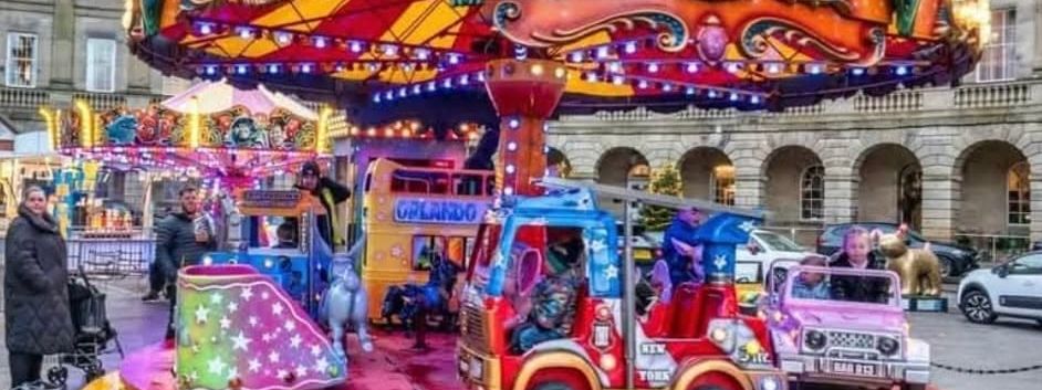 The large juvenile toy set roundabout on a wet paved town square, with the yellow Orlando double-decker bus, red fire engine and pink jeep on the bulb-ringed deck under the orange glitter canopy