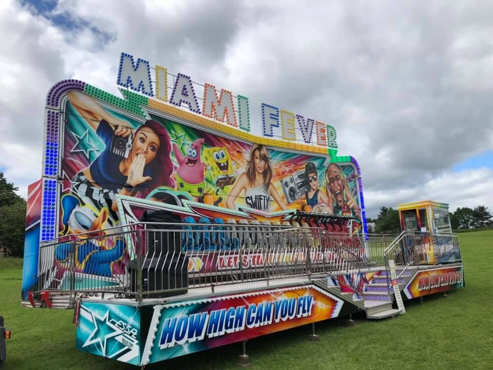 Full Miami Fever ride set up on grass beneath a cloudy sky, with its painted frontage, loading platform and control booth