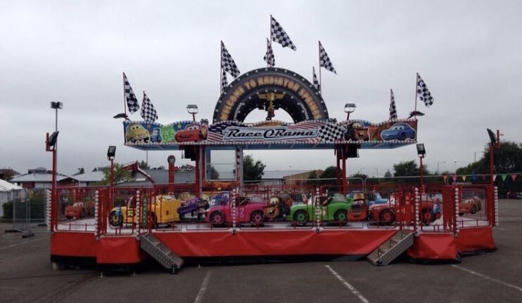 Race-a-Rama ride set up on tarmac with character cars on the platform, checkered flags, tyre-arch sign and central operator booth