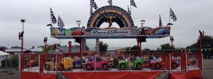 Race-a-Rama ride set up on tarmac, with character cars on a circular platform, checkered flags along the roofline and a tyre-arch sign above the painted frontage