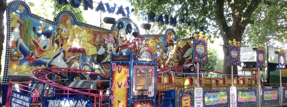 Runaway Train funfair ride set up on tarmac beneath trees, with its painted wild-west frontboard, blue star-decked lettering and fenced track