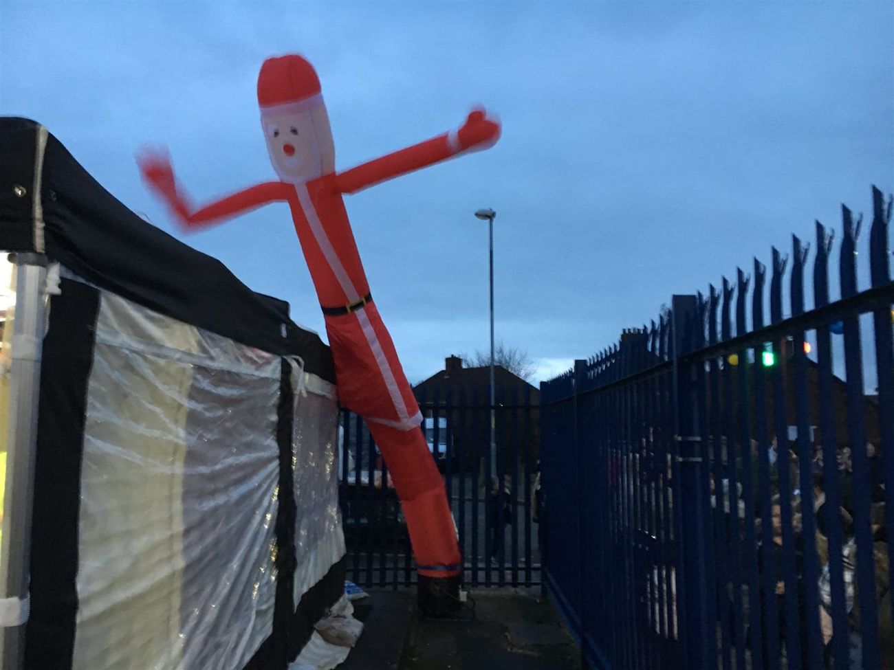 The Santa sky dancer leaning dramatically beside a marquee tent at dusk, showing the full range of arm motion