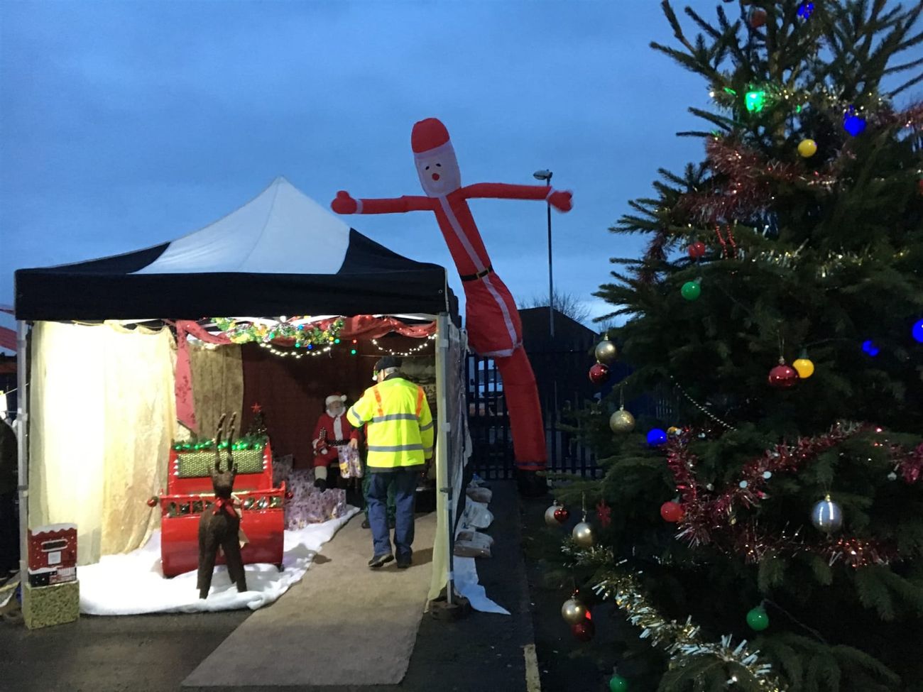 The Santa sky dancer towering over a Christmas grotto marquee at dusk, with a decorated tree and festive string lighting