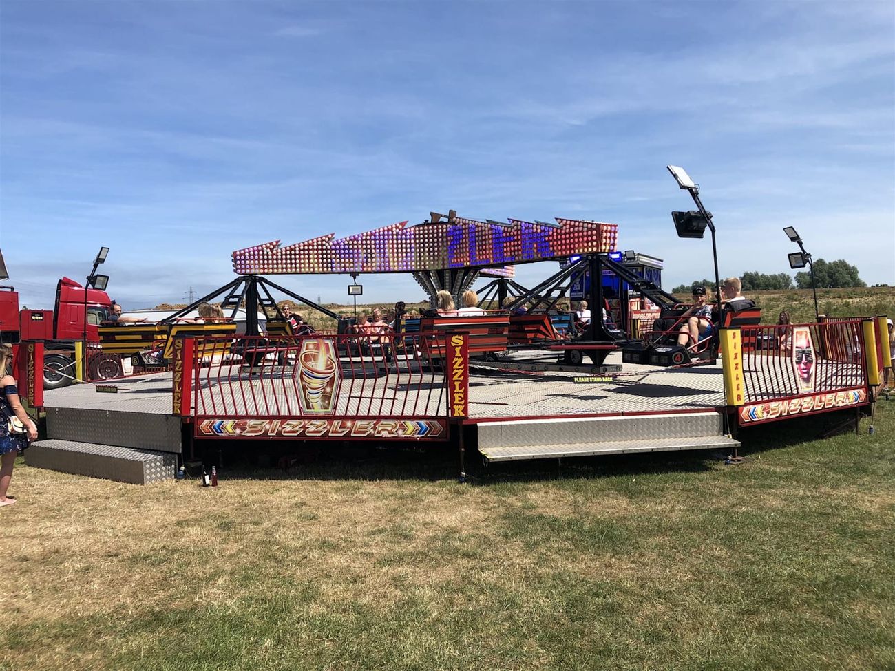 Twister Sizzler ride operating on grass with riders in red and yellow bench cars around the illuminated centre