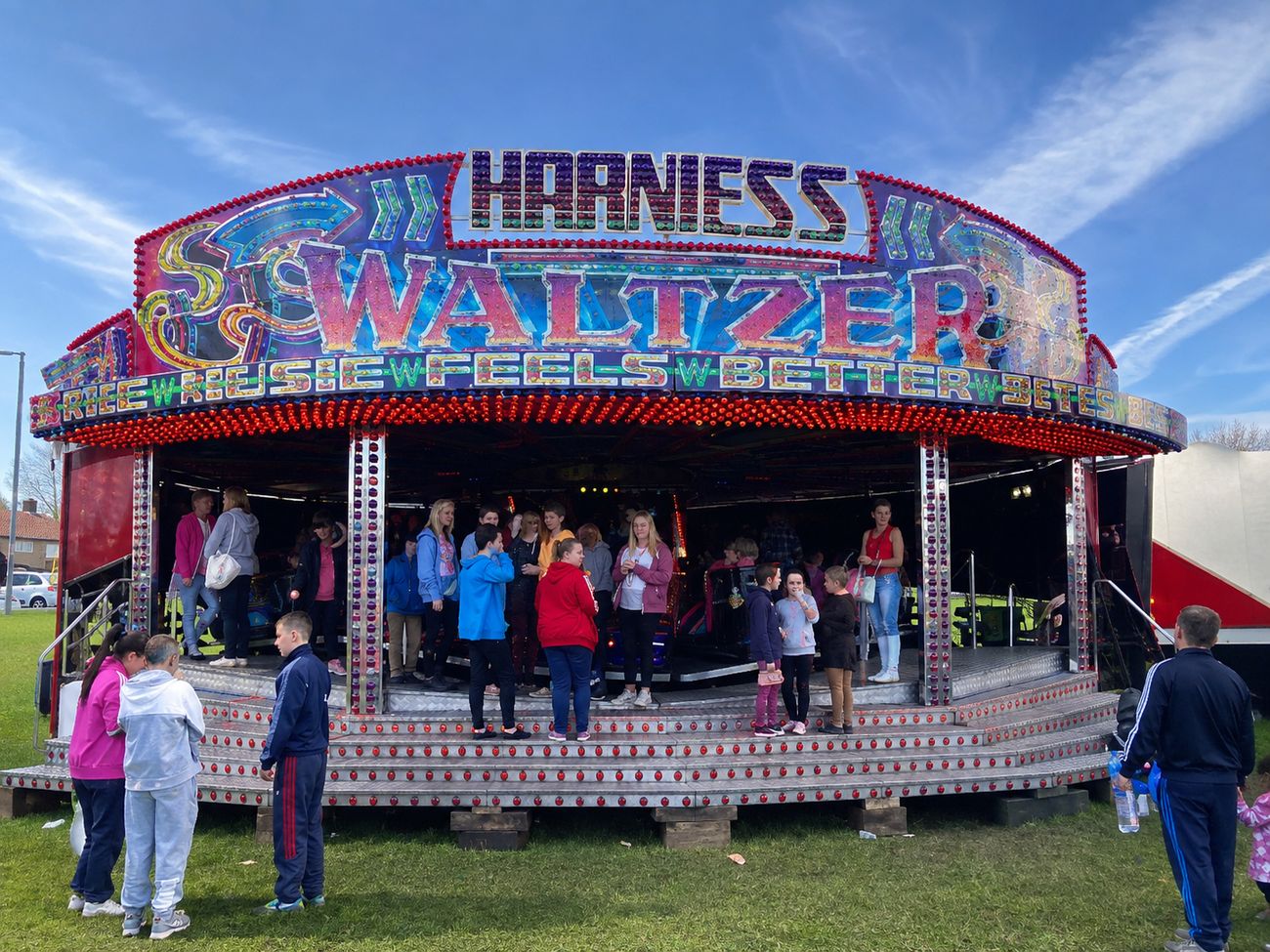 Harniess Waltzer set up on grass in daylight, with its full illuminated sign and a crowd gathered across the stepped loading deck